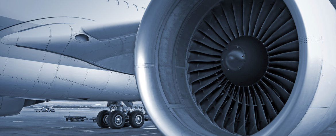 Nahaufnahme eines an einem Verkehrsflugzeug montierten Turbofan-Strahltriebwerks, stationär, mit sichtbaren Lüfterschaufeln; angrenzendes Fahrwerk und Flughafenvorfeld mit kleinen Bodenfahrzeugen im Hintergrund.
