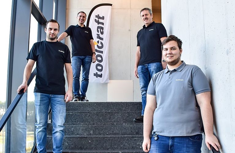 Four men stand on a concrete stairwell, smiling and leaning on a railing by large windows; a vertical banner behind them reads "toolcraft" and their shirts display "toolcraft".
