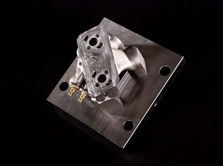 A precision-machined metal engine component sits secured on a rectangular steel mold plate, its milled surfaces, two brass fittings, and circular ports visible under studio lighting against a black background.