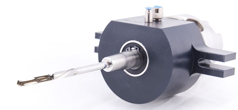 A metal twist drill bit mounted in a black cylindrical spindle assembly projects forward, stationary and centered in a studio-lit close-up on a white background.