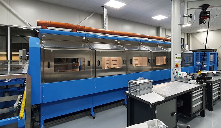 A long blue industrial processing machine with multiple windowed compartments sits idle on a clean factory floor, flanked by workbenches, overhead orange piping, and bright ceiling lights.
Visible text: 7 6 5