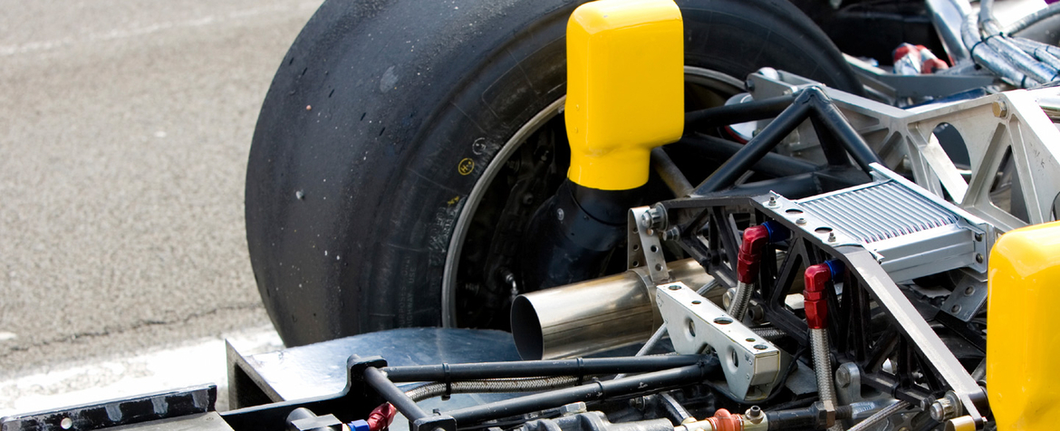 Hinterrad und freiliegende Fahrwerkskomponenten des Rennwagens – Reifen, Auspuffrohr, Aufhängungsstreben und Ladeluftkühler – stehen still auf Asphalt in einer Boxengasse/Rennstrecke; gelbe Crashblöcke sind sichtbar.