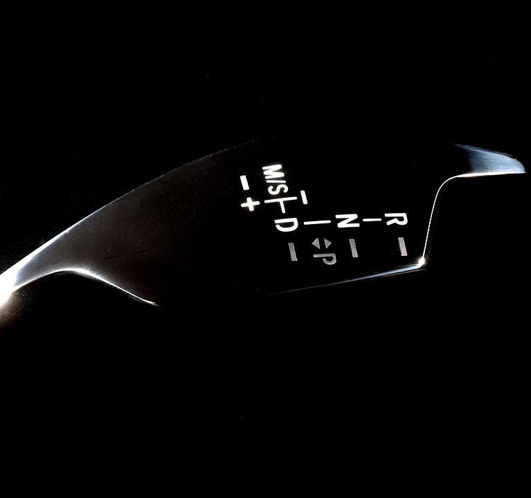 Car gear-selector panel displays illuminated gear positions against a deep black background, reflective edges catching highlights.

Text transcription (as printed): R  N  D  P  (left-right arrow symbol)  M/S  -  +