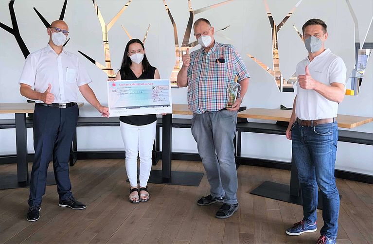 Ein übergroßer Bankscheck wird von vier maskierten Erwachsenen mit erhobenem Daumen in einem hellen Raum mit weißen, baumförmigen Wandausschnitten gehalten.
Transkribierter Text sichtbar:
- Sparkasse Mittelfranken-Süd
- 3.000,- €
Weitere handschriftliche Angaben, Unterschriften, Konto-/IBAN-Nummern und kleinere Druckbuchstaben sind auf dem Scheck vorhanden, aber auf dem Bild nicht lesbar.