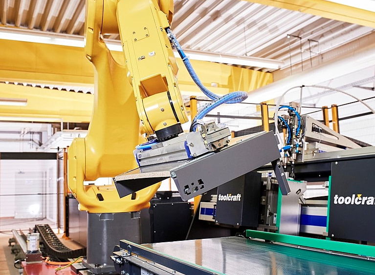 Yellow industrial robotic arm grips a gray sheet-metal gripper, positioning it above a conveyor table amid machines labeled "toolcraft" inside a brightly lit factory workshop.
