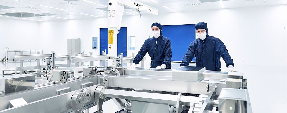 Heller Reinraum mit mehreren Anlagen zur technischen Feinstreinigung von Bauteilen, zwei Personen in reinraumkleidung schieben einen Wagen.