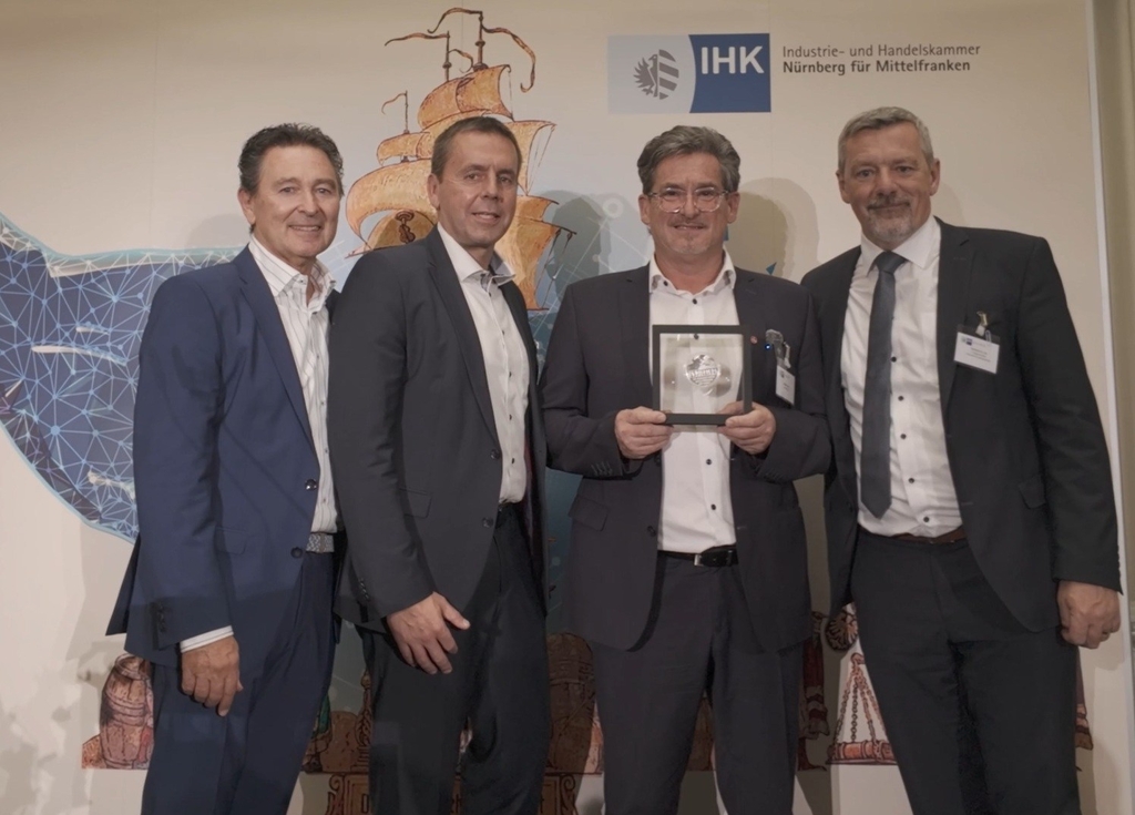 Ein Mann (Mitte rechts) hält eine gerahmte Auszeichnung in der Hand, flankiert von drei lächelnden Männern im Anzug vor einer Kulisse mit der Aufschrift „IHK Industrie- und Handelskammer Nürnberg für Mittelfranken“.