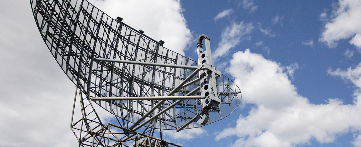 Large open-frame metal radar dish tilting upward, its lattice supports and feed arm visible against a partly cloudy blue sky.