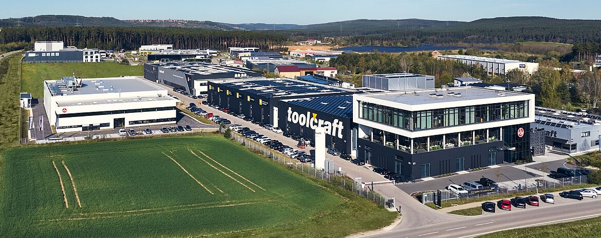 Ein Industriekomplex aus schwarz-weißen Fabrikgebäuden der Firma „Toolcraft“ liegt neben geparkten Autos und einer Rasenfläche in einem Gewerbegebiet mit umliegenden Lagerhallen und Bäumen. Auf mehreren Gebäuden ist der Schriftzug „Toolcraft“ zu sehen.