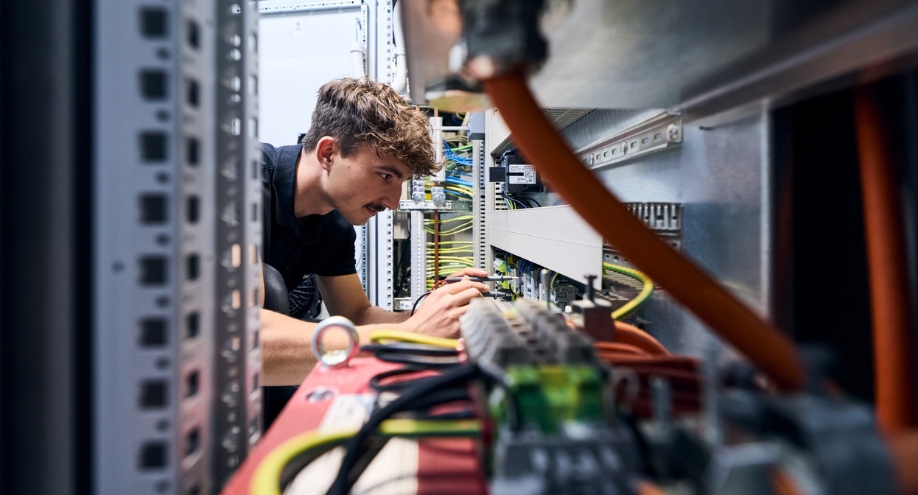 Ein Techniker prüft mit Multimeterleitungen die Klemmenblöcke eines elektrischen Schaltschranks. Sie sind von gebündelten grünen, schwarzen und blauen Kabeln umgeben. Text: Kleine weiße Klemmenbeschriftungen nicht lesbar.