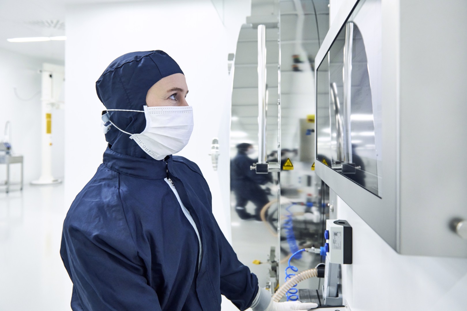 Heller Reinraum mit mehreren Anlagen zur technischen Feinstreinigung von Bauteilen, einschließlich Reinigungsanlagen, Fördertechnik und automatisierter Handhabungssysteme. Vorne steht eine Frau im Profil in einem blauen Schutzanzug mit einer weißen Maske und blickt in einen Bildschirm.